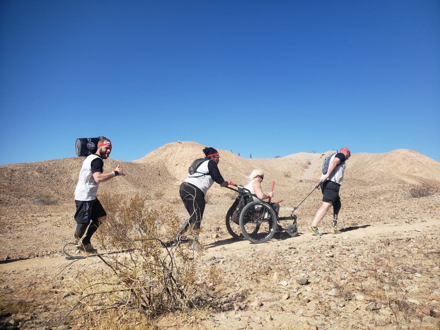 Three people walk alongside a fourth person in a wheelchair on a desert trail among dunes.